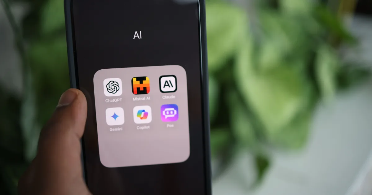 Hand holding a smartphone showing a folder of AI assistant apps, including ChatGPT, Claude, and Gemini